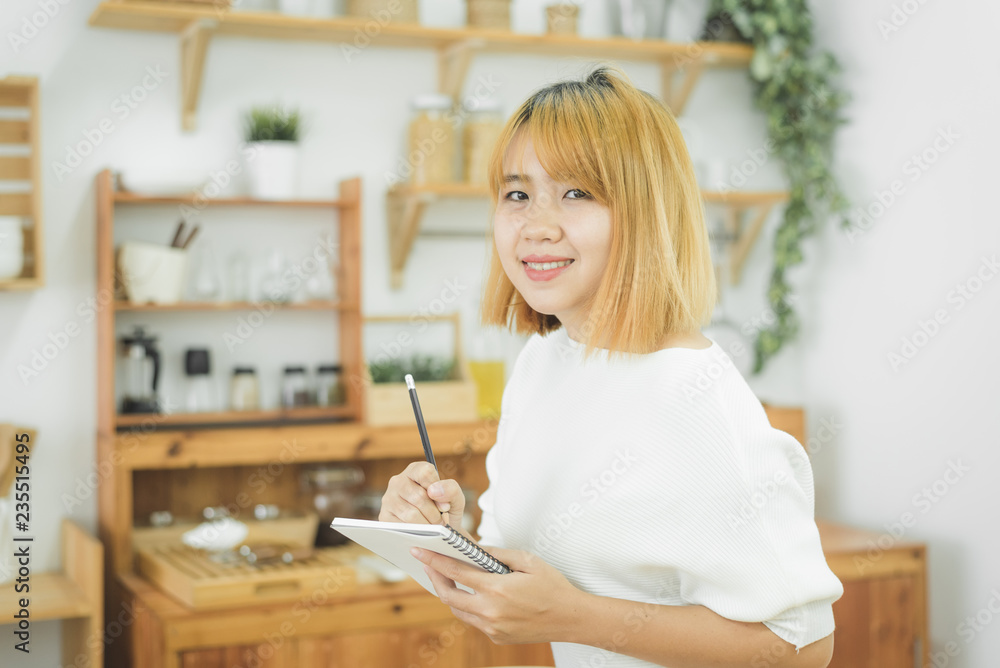 Asian woman write shopping lists in notepad by pen on her kitchen counter at home and reading and tries to remember all ingredients to shopping in grocery. Lifestyle women relax at home concept. Asian woman write shopping lists in notepad by pen on her kitchen counter at home and reading and tries to remember all ingredients to shopping in grocery. Lifestyle women relax at home concept.