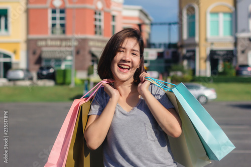 Happy young Asian woman shopping an outdoor market with background of pastel buildings and blue sky. Young asian woman smile with a colorful bag in her hand. Outdoor woman lifestyle shopping concept. © tirachard