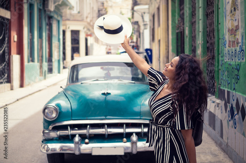 Young beautiful black woman with a typical cuban hat standing in the old streets of Havana Cuba in front of a classic car holding the hat right over the car. © Saga_bear
