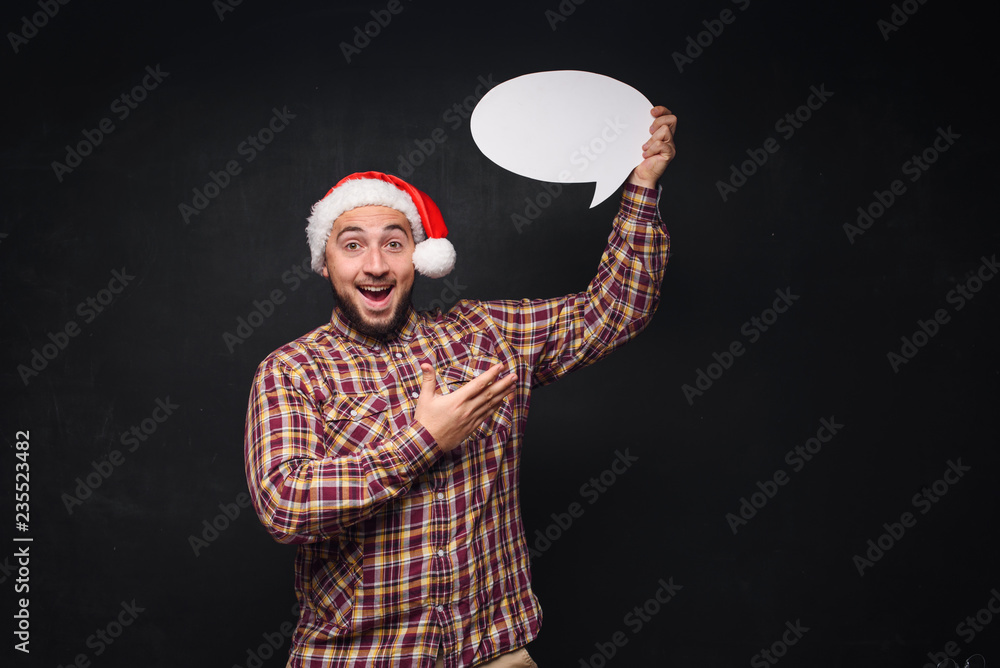 Funny serious man in red Christmas Santa hat holds empty white cardboard as blank or mock up with copy space for text. Black background Funny serious man in red Christmas Santa hat holds empty white cardboard as blank or mock up with copy space for text. Black background