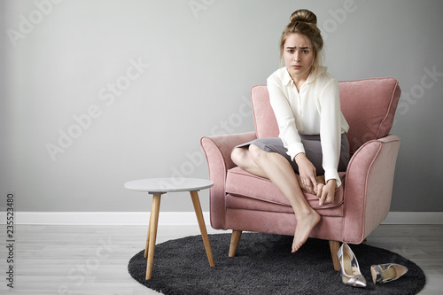 Billede på lærred Modern hardworking young female feeling tired after long walk on high heeled shoes, sitting barefooted in armchair, massaging her feet, having painful frustrated facial expression