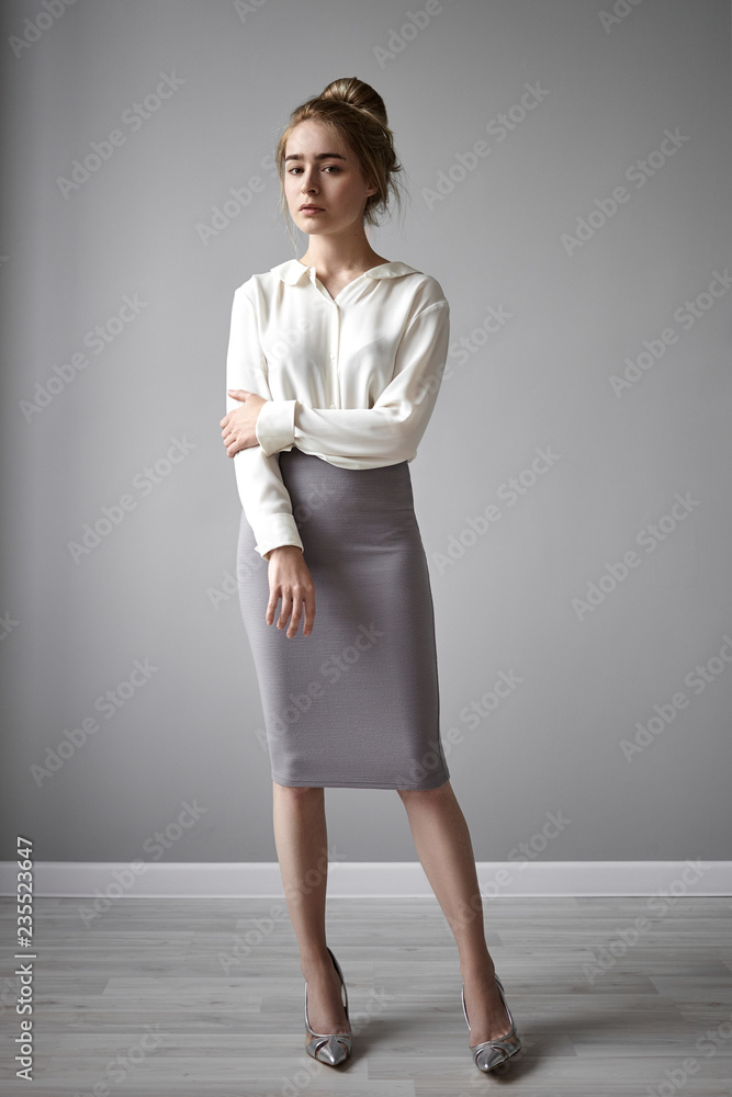 Vertical picture of gorgeous stylish young European lady in elegant white  blouse and tube skirt holding hand on arm, staring at camera with serious  confident look. Style, beauty, glamour and fashion Photos