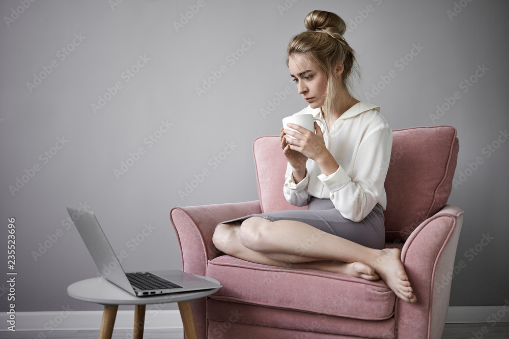 Picture of attractive barefooted young blonde Caucasian woman dressed in skirt and white blouse having morning coffee from larger mug while watching serious online on generic notebook computer