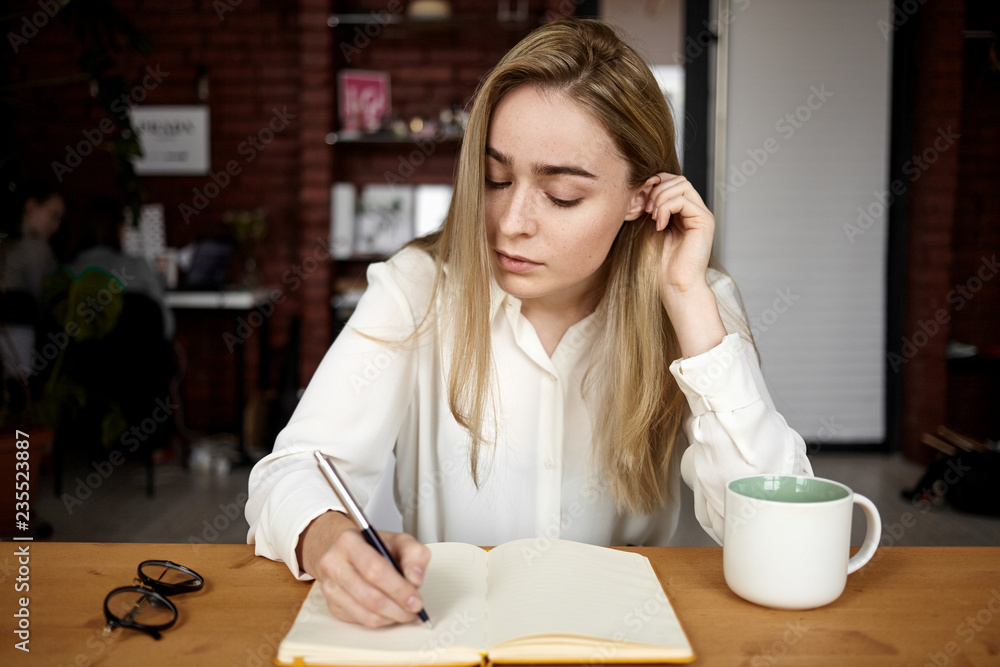 Candid shot of attractive blonde student girl in white blouse doing homework at workplace at home, writing down in open copybook, drinking tea, having serious concentrated facial expression Candid shot of attractive blonde student girl in white blouse doing homework at workplace at home, writing down in open copybook, drinking tea, having serious concentrated facial expression