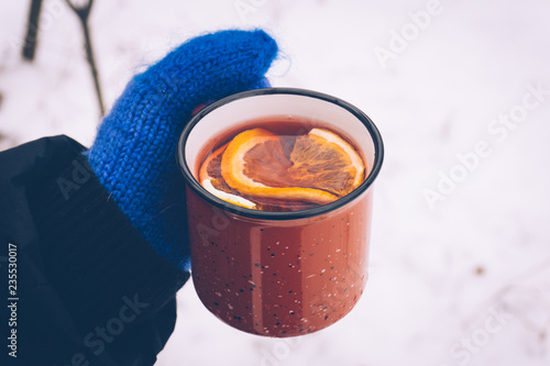 Mulled wine in a red cup in women's hands in mittens. Hot winter drink outdoor in snowy winter forest in cold weather. Christmas background with snowflakes