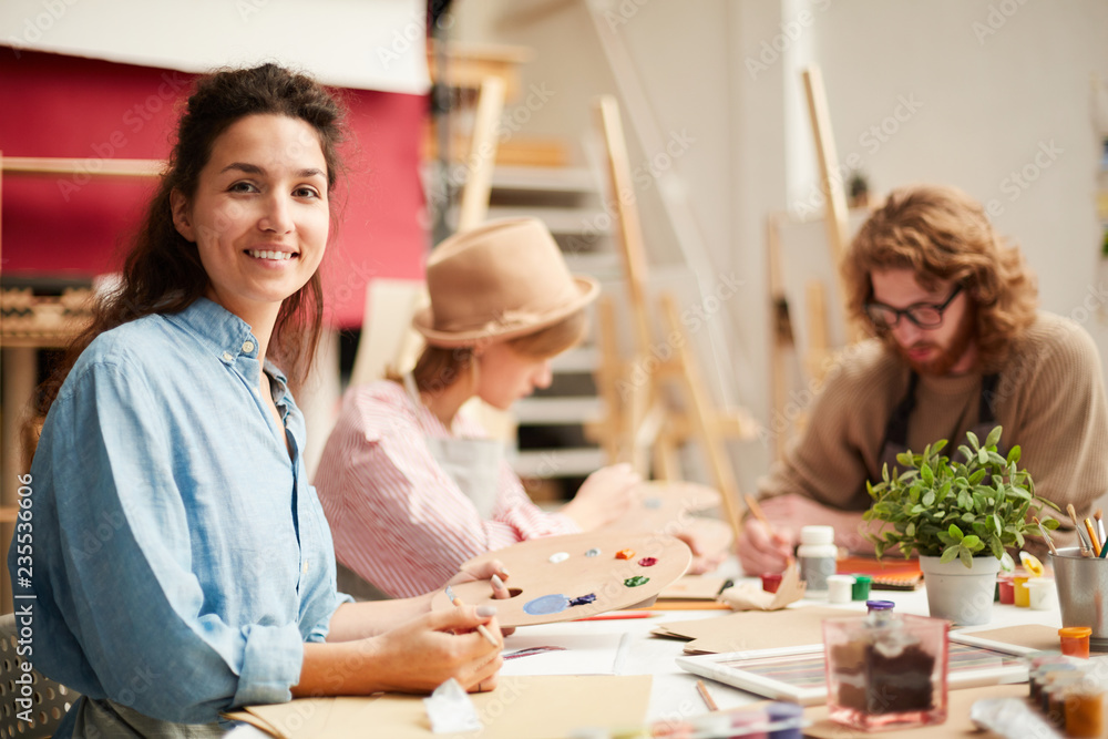 One of students with palette looking at you while sitting by table on background of groupmates One of students with palette looking at you while sitting by table on background of groupmates