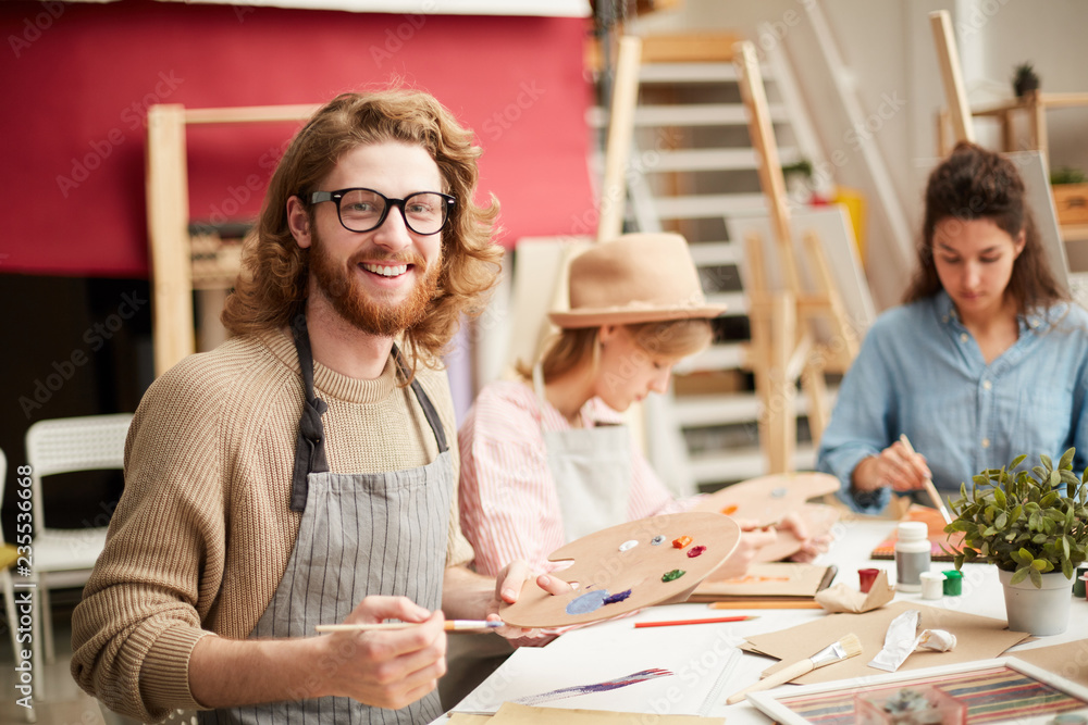 Happy young bearded man in apron looking at camera while doing painting work by table Happy young bearded man in apron looking at camera while doing painting work by table