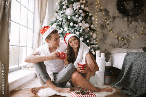Happy guy and girl in white t-shirts and Santa Claus hats sit with red cups on the floor in front of the window next to the New Year tree, gifts and candles