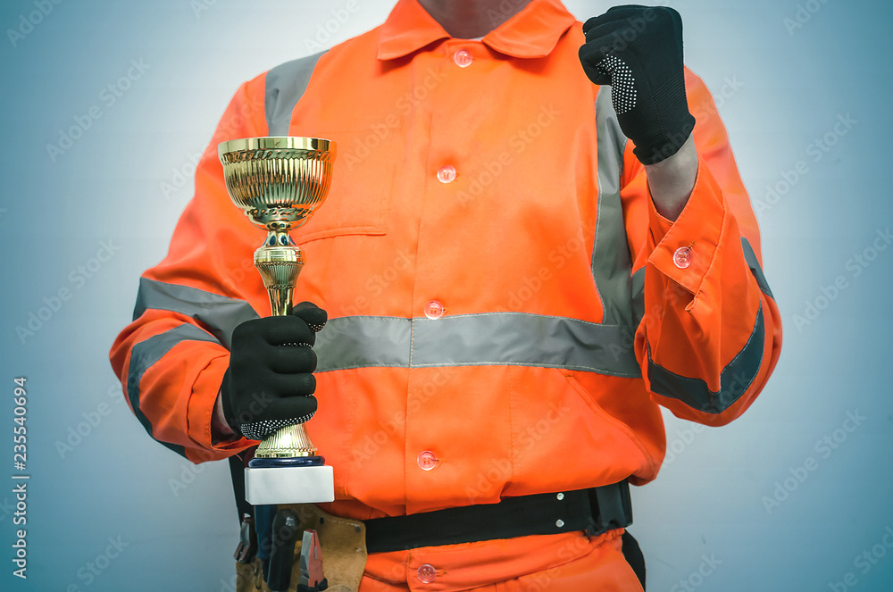 Best builder contractor award concept. Golden cup trophy in the construction worker hand isolated on blue background. Best builder contractor award concept. Golden cup trophy in the construction worker hand isolated on blue background.