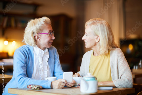 Two mature blonde women sitting by table in cafe and discussing online news by cup of tea