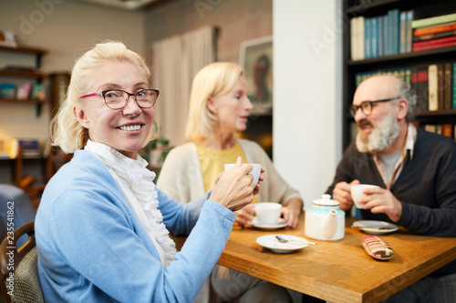 Happy blonde aged female with cup of tea sitting by table with her senior friends in cafe