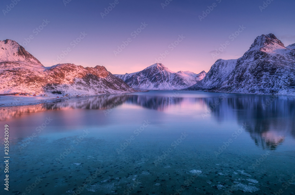 Aerial view of snow covered mountains and colorful sky reflected in water at dusk. Winter landscape with sea, snowy rocks, purple sky, reflection at sunset. Lofoten islands, Norway at twilight. Nature