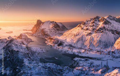 Aerial view of beautiful fjord at sunset in Lofoten Islands, Norway. Winter landscape with snowy mountains, blue sea and orange sky with sun. Top view of rocks in snow, road, village. North coastline