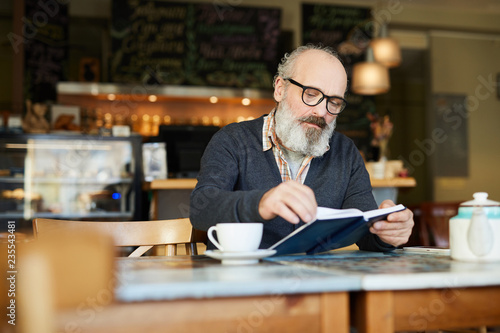 Senior bearded man sitting in cafe and turning page of notebook or book while reading it by cup of tea