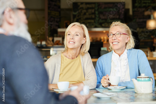 Two pretty aged cheerful women in smart casual looking at senior man during their talk in cafe