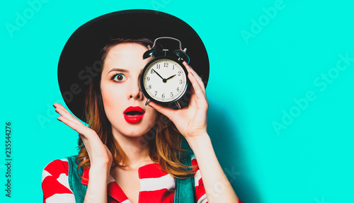 Portrait of young suprised red-haired white european woman in hat and red striped shirt with alarm clock on blue background