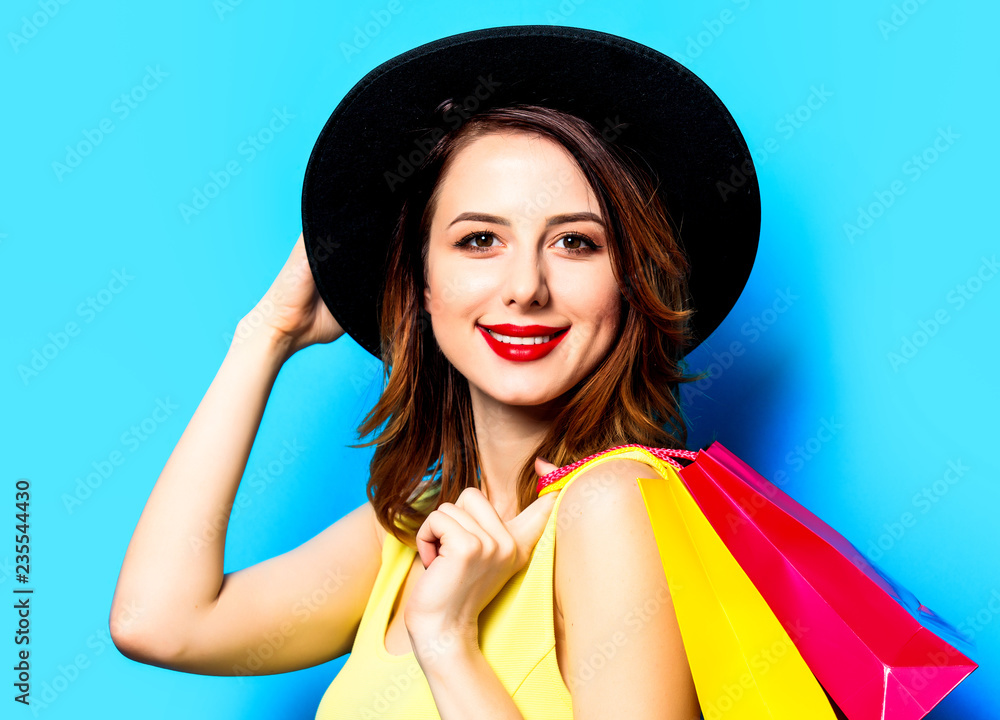 Fototapeta premium Portrait of young smiling red-haired white european woman in hat with shopping bags on blue background
