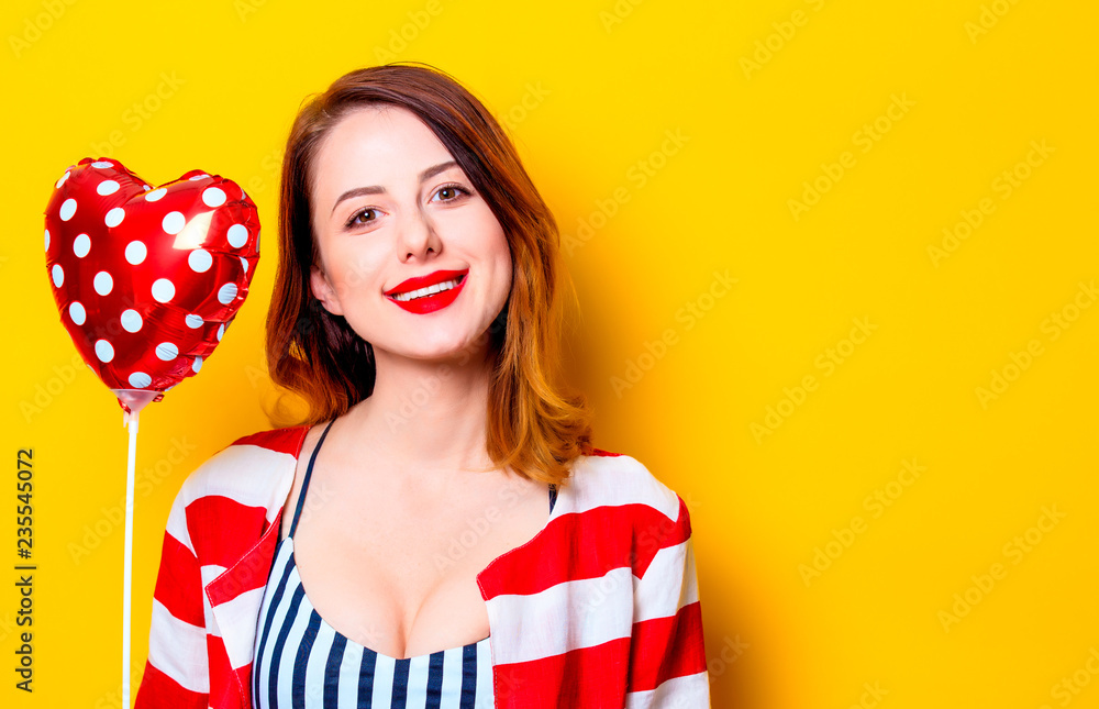 Portrait of beautiful young smiling red-haired white european woman in red striped shirt with heart shape toy on yellow background Portrait of beautiful young smiling red-haired white european woman in red striped shirt with heart shape toy on yellow background