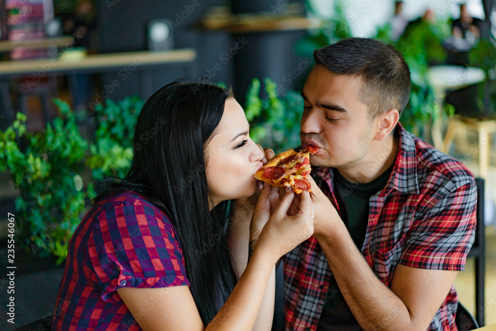 A pretty young couple eating one slice of pizza together. A nice girl and handsome man biting the same pizza in a modern cafe. A pretty young couple eating one slice of pizza together. A nice girl and handsome man biting the same pizza in a modern cafe.