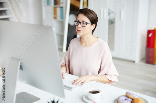 Young elegant businesswoman looking at computer screen while networking by desk in office