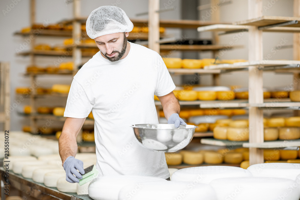Fototapeta Mężczyzna tarcie kół serowych woskiem w produkcji sera z półkami pełnymi sera w tle