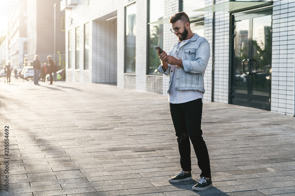 Handsome smiling hipster businessman with beard, in denim jacket, trendy glasses walks around city and using smartphone. Handsome smiling hipster businessman with beard, in denim jacket, trendy glasses walks around city and using smartphone.