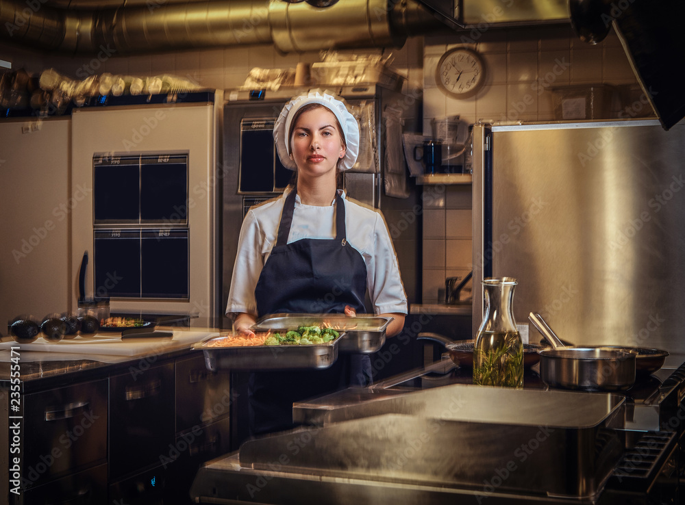 Female chef wearing a uniform holding a container with chopped vegetables at restaurant's kitchen. Female chef wearing a uniform holding a container with chopped vegetables at restaurant's kitchen.