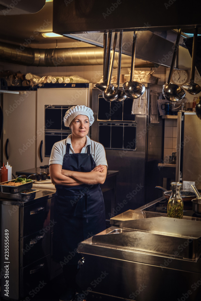 A middle age cook wearing a uniform standing with her arms crossed at restaurant's kitchen. A middle age cook wearing a uniform standing with her arms crossed at restaurant's kitchen.