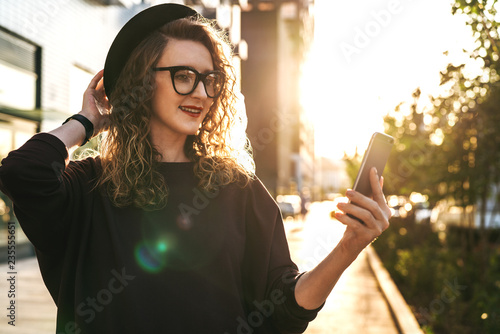 Young curly woman in hat and fashionable glasses makes photo on digital camera of smartphone  standing on city street.