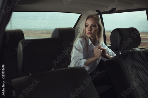 Portrait of beautiful woman sitting on the back seat of the old car and looking at the window thoughtfully. Drops of rain outside on the back window. Movie shot concept. Text space
