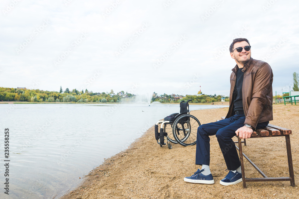 Obraz premium A young man sits on a bench by the lake, next to his wheelchair.