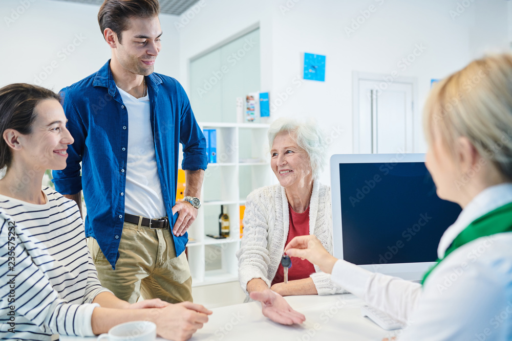 Obraz premium Positive adult children taking mortgage for mother in bank while making present for her: bank manager giving flat key to senior lady thanking son and daughter