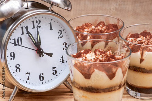Diet concept with alarm clock: sweet Italian dessert tiramasu with delicate mascarpone cream from traditional recipe in glass on wooden background