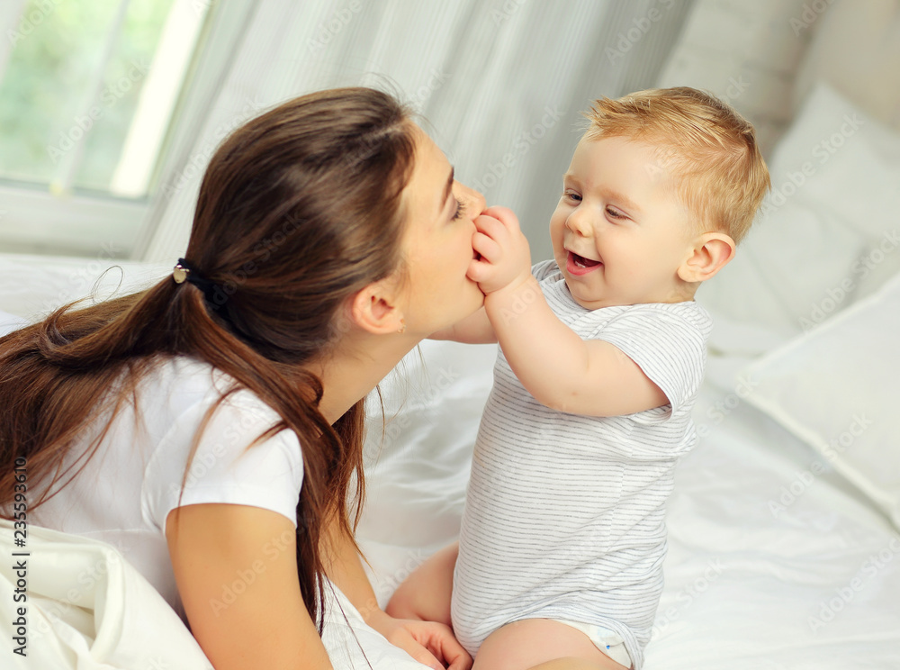 Mom and a charming blue-eyed baby of 10 months in a striped jumpsuit are lying in bed and hugging Mom and a charming blue-eyed baby of 10 months in a striped jumpsuit are lying in bed and hugging
