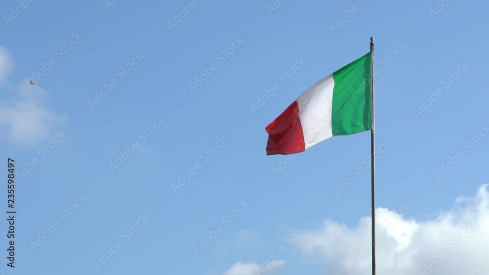 Italian flag waving in wind on flagpole with a seagull flight in background at an Italy city. Italian banner flutters with bird flying at day with blue sky. Real fabric texture ensign fluttering-Dan Italian flag waving in wind on flagpole with a seagull flight in background at an Italy city. Italian banner flutters with bird flying at day with blue sky. Real fabric texture ensign fluttering-Dan
