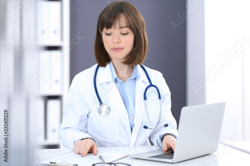 Doctor typing on laptop computer while sitting at the white table in hospital office. Physician at work. Medicine and healthcare concept