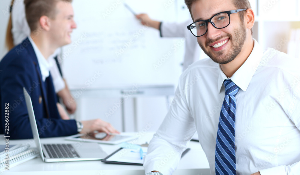 Business people at meeting in office. Focus at cheerful smiling bearded man wearing glasses. Conference, corporate training or brainstorming of people group. Success and negotiation concept Business people at meeting in office. Focus at cheerful smiling bearded man wearing glasses. Conference, corporate training or brainstorming of people group. Success and negotiation concept