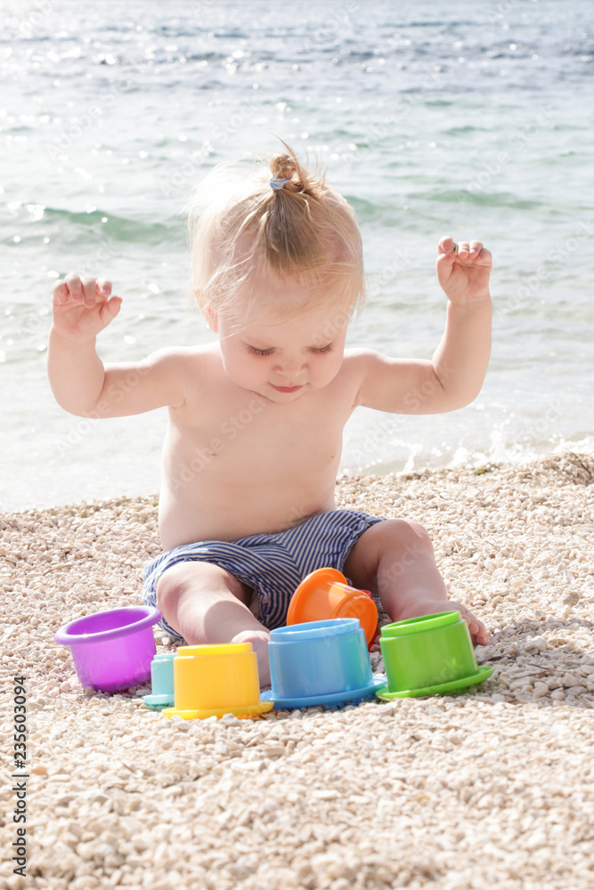 Cute baby playing with plastic toys on the pebble beach near the sea Cute baby playing with plastic toys on the pebble beach near the sea