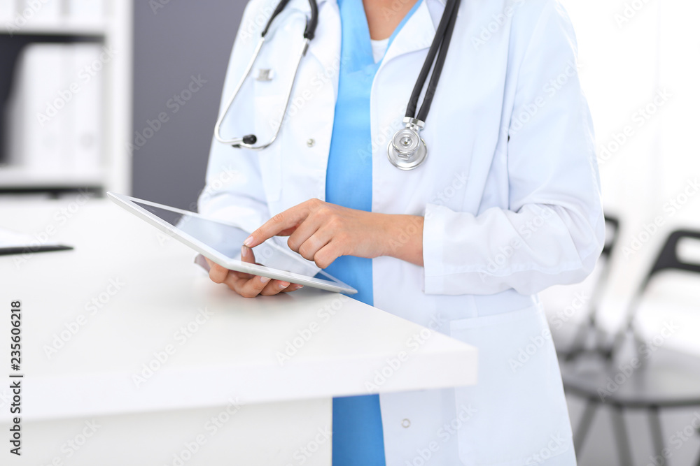 Close-up of female physician using digital tablet while standing near reception desk at clinic or emergency hospital. Unknown doctor woman at work. Medicine and healthcare concept Close-up of female physician using digital tablet while standing near reception desk at clinic or emergency hospital. Unknown doctor woman at work. Medicine and healthcare concept