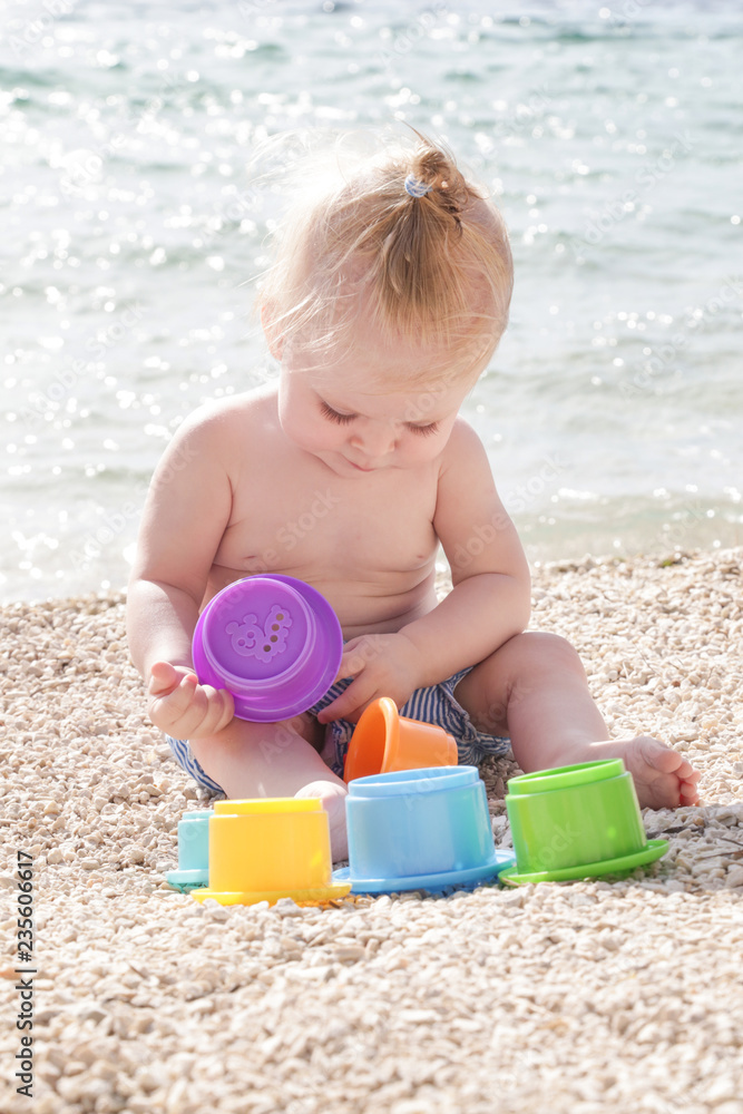Cute baby playing with plastic toys on the pebble beach near the sea Cute baby playing with plastic toys on the pebble beach near the sea