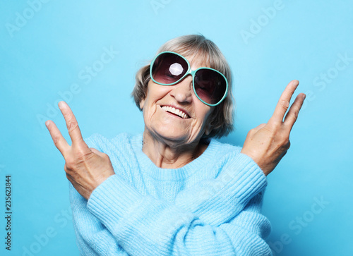 Funny old lady wearing blue sweater, hat and sunglasses showing victory sign.