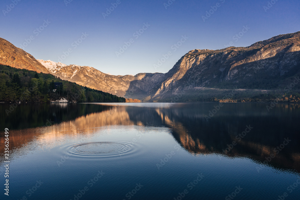 Sunrise view of alpine lake Bohinj with blue sky, sunlight and mist over water. Circles in water. Famous lake of Bohinj in Triglav National Park, Julian alps, Slovenia. Sunrise view of alpine lake Bohinj with blue sky, sunlight and mist over water. Circles in water. Famous lake of Bohinj in Triglav National Park, Julian alps, Slovenia.