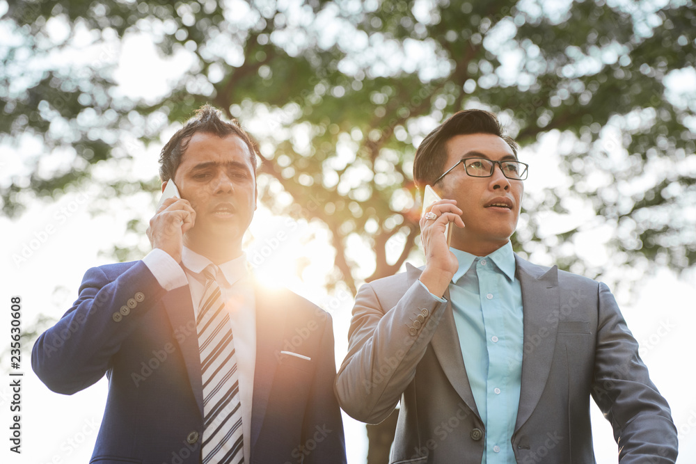 Two businessmen in formal suits standing outdoors and talking on mobile phones Two businessmen in formal suits standing outdoors and talking on mobile phones