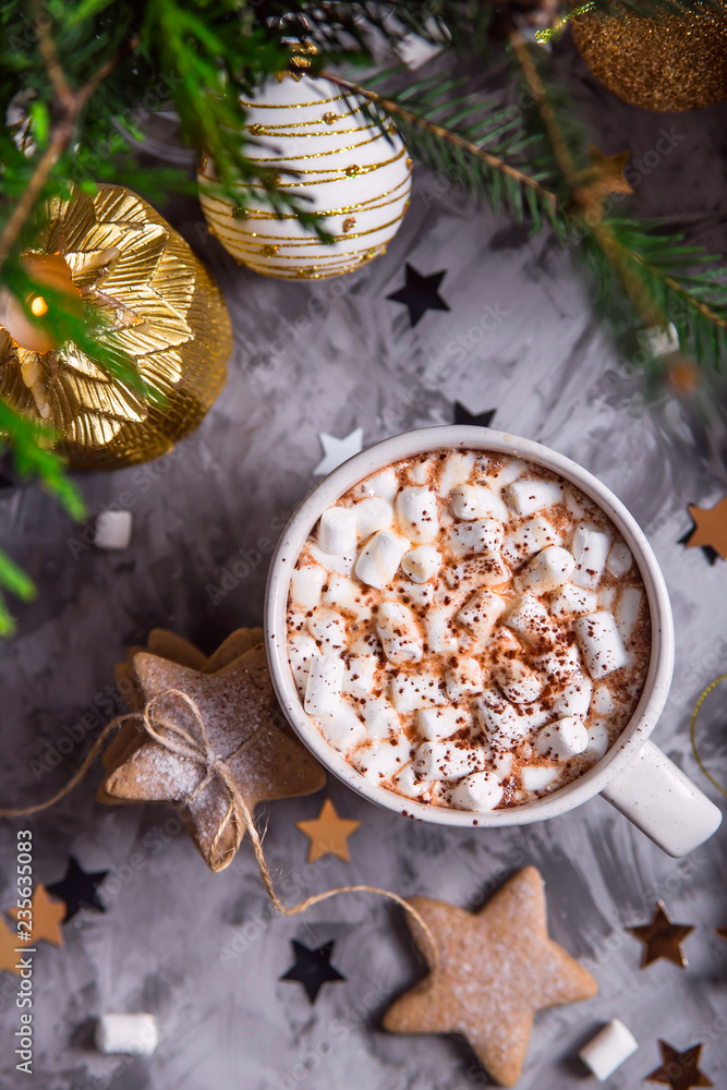A large cup of cocoa with marshmallow sprinkled with cocoa powder stands on a gray table among Christmas decorations, fir branches, ginger cookies and shiny stars. Top view A large cup of cocoa with marshmallow sprinkled with cocoa powder stands on a gray table among Christmas decorations, fir branches, ginger cookies and shiny stars. Top view