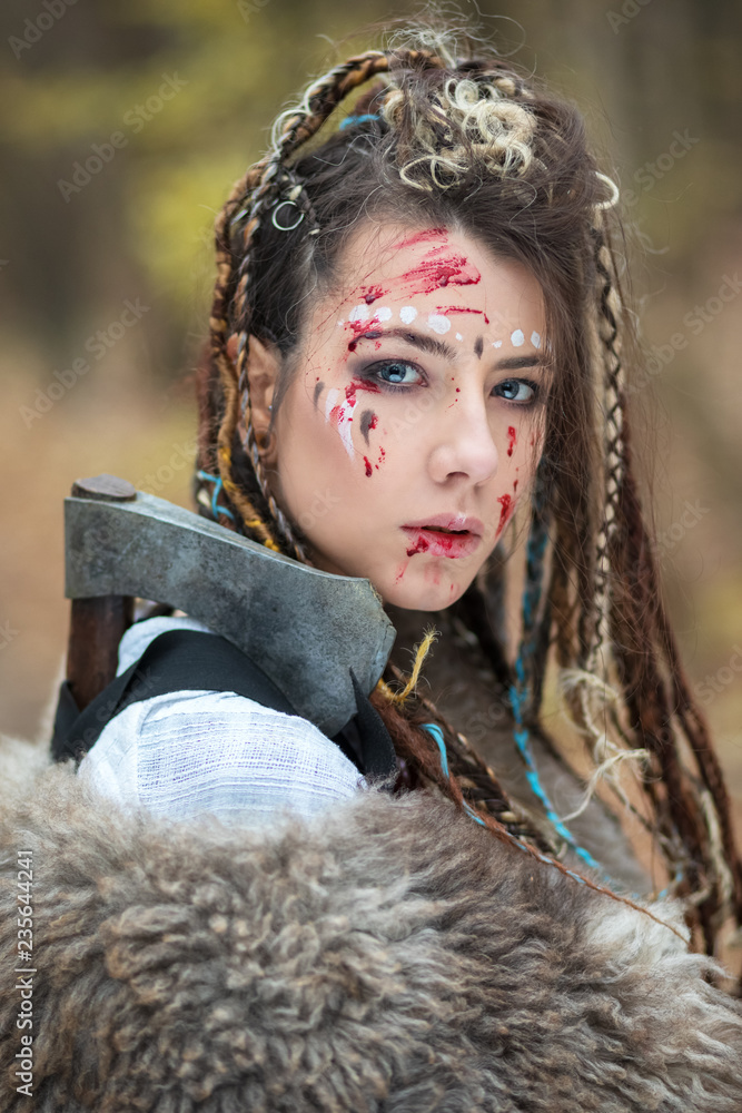 Foto Stock Closeup portrait of Viking warrior woman with war makeup and  braided hair with face covered in blood after battle, ready to attack.  Northern warrior woman alone in forest with ax.