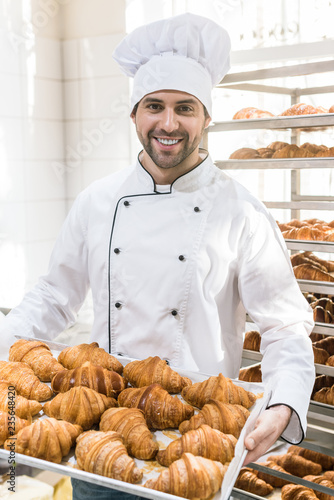 Smiling baker in white chefs uniform with tray full of fresh croissants