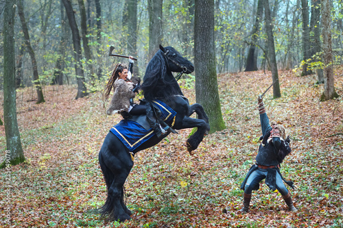 Portrait of dangerous scandinavian viking woman riding black horse in forest holding ax fighting with Viking warrior man in forest.