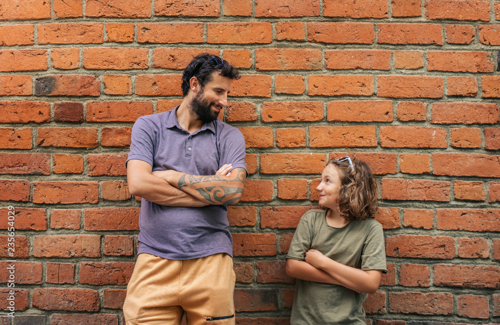 Content son and father leaning against a brick wall outside - obrazy, fototapety, plakaty 