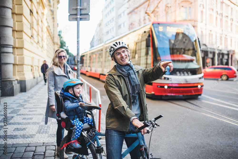 Obraz premium A small toddler boy sitting in bicycle seat with young parents outdoors in city.