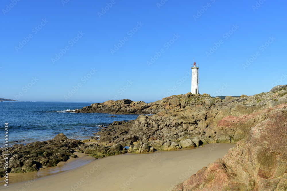 Beach with rocks and white lighthouse. Wet sand, blue sea, sunny day. Galicia, Spain.